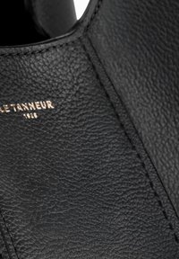 Schwarzes Leder mit strukturierter Oberfläche. Prägung in Gold mit der Marke "LE TANNEUR" und dem Jahr "1880". Nahaufnahme der Nahtdetails.