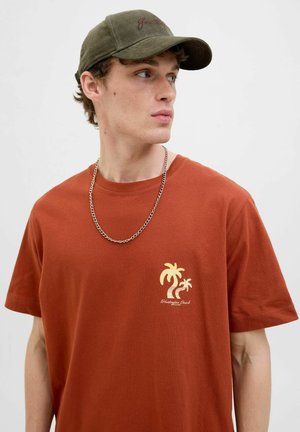 Camiseta de algodón marrón con un gráfico de palmera dorada en el lado izquierdo del pecho. Combinada con una gorra verde oscuro y un collar de cadena plateada.