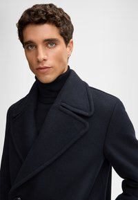 Navyblauer Wollmischmantel mit spitzen Revers, doppelter Knopfleiste und weicher Textur. Über einem schwarzen Rollkragenpullover getragen.