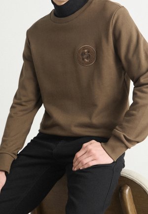Person iført en brun sweater med et cirkulært emblem og sorte bukser, siddende med hænderne hvilende på benene mod en ensfarvet baggrund.