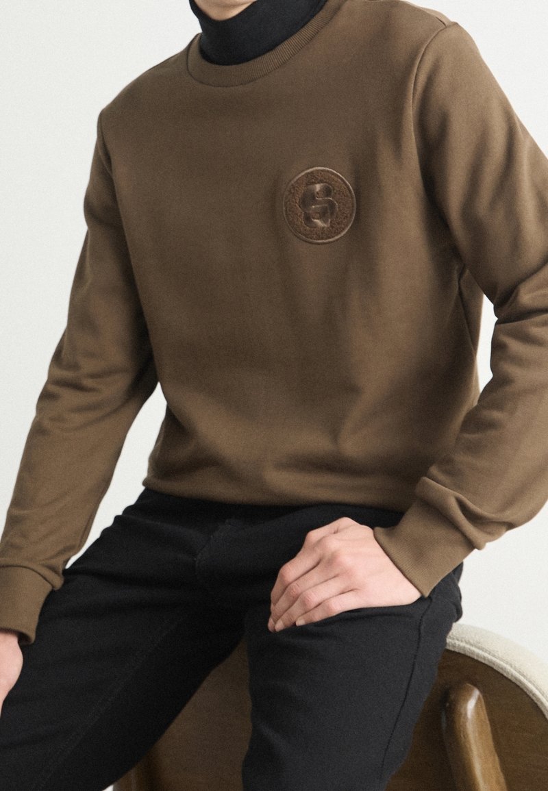 Personne portant un pull marron avec un emblème circulaire et un pantalon noir, assise les mains posées sur les jambes devant un fond uni.