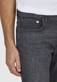 Ciemnoszare dżinsy slim fit o gładkiej fakturze, z detalami kieszeni z przodu oraz metalowymi elementami do zapięcia, pokazane obok ręki.