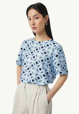 Light blue blouse featuring a geometric pattern in navy and white. Round neckline, short sleeves, and a relaxed fit. Smooth fabric texture.