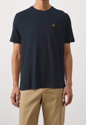 Mann trägt ein marineblaues, kurzärmeliges, geripptes T-Shirt mit Brusttasche und kleinem gelben Logo, kombiniert mit beigen Hosen.