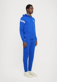 Blue zip-up hoodie with side pockets and zip closure, paired with matching joggers; features white striped accents and textured fabric.