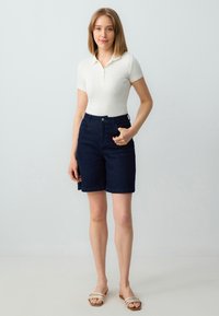 Witte geribde polo, donkerblauwe denim shorts, korte zoom, voorzakken en lichtgekleurde slidersandalen met meerdere banden.
