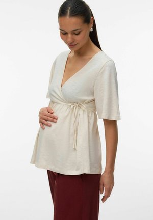 Rase naine tume juustega beežis mähistopis ja burgundia värvi pükstes, hoides õrnalt oma kõhtu ja vaadates alla pehme naeratusega.