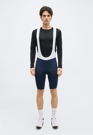 Fiatal férfi áll, fekete hosszú ujjú felsőt, sötétkék kerékpáros nadrágtartós rövidnadrágot fehér pántokkal, fehér zoknit és fehér kerékpáros cipőt viselve, egyszínű háttér előtt.