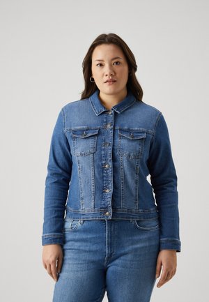 Femme aux longs cheveux foncés portant une veste en jean bleue boutonnée et un jean bleu assorti, debout devant un fond clair uni.