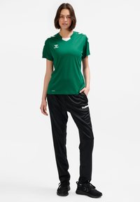 Grünes Sporttrikot mit kurzen Ärmeln, V-Ausschnitt und weißen Akzenten, kombiniert mit schwarzen, locker sitzenden Trainingshosen. Beide aus synthetischen Materialien gefertigt.