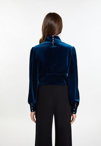 Samt-Bluse mit hohem Kragen in einem tiefen Blau, mit puffärmel, Knopfdetails an den Manschetten und einem Rückenschlitz mit dekorativen Knöpfen.