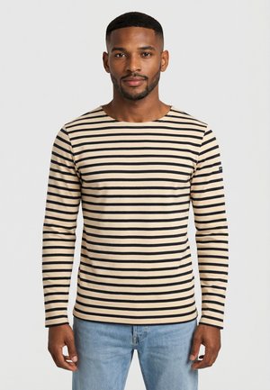 Homme portant une chemise à manches longues à rayures horizontales beiges et noires, et un jean bleu clair, debout devant un fond uni.