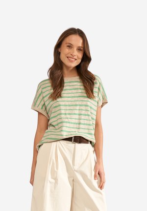 Vrouw glimlachend, draagt een beige en groen gestreept kortärmelig topje in een wit broek met hoge taille en een bruine riem, staand tegen een witte achtergrond.