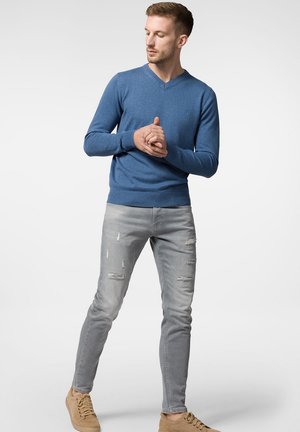 Niebieski sweter w serek z prążkowanymi mankietami, zestawiony z jasnoszarymi, zniszczonymi jeansami skinny i beżowymi sneakerami. Prosty i nowoczesny design.