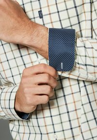 Chemise à carreaux avec des motifs crème, vert et rouille. Manches contrastées en marine avec un design à pois. Fermeture à bouton sur les manches et le collier.