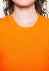 Haut en maille côtelée orange avec un col rond et un détail texturé diagonal sur le devant. Matière douce et extensible avec une finition lisse.