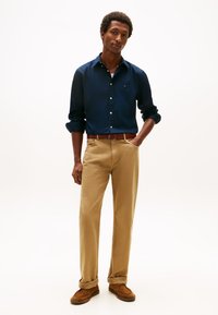 Marineblauw overhemd met knopen, beige chino's, bruine riem en beige schoenen. De outfit heeft een klassieke pasvorm en opgerolde mouwen aan het overhemd.