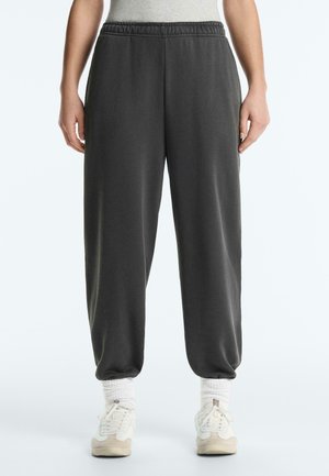 Osoba ubrana w ciemnoszare dresowe spodnie z elastycznym pasem, białe skarpetki i białe trampki na jednolitym jasnym tle.