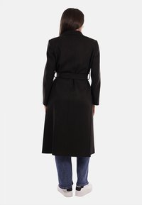 Donna in piedi di spalle con un lungo cappotto nero con cintura, jeans blu e scarpe bianche su uno sfondo bianco uniforme.