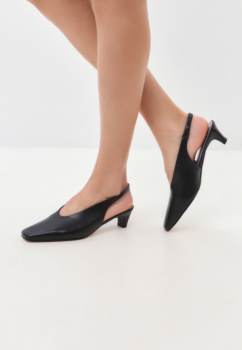 Zapatos de piel negros tipo slingback con punta afilada, tacón bajo y laterales con cortes. Textura suave y diseño elegante.