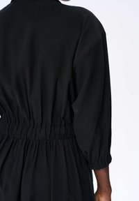 Czarny kombinezon z elastycznym paskiem w talii, posiadający długie rękawy i subtelne marszczenia. Gładka tekstura materiału. Brak widocznych wzorów i elementów metalowych.