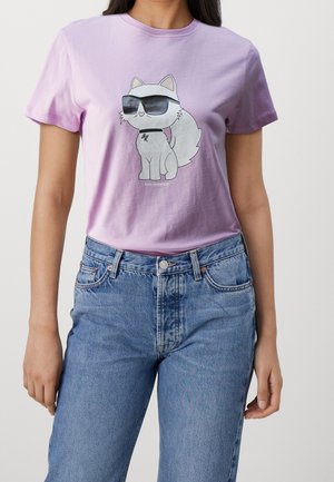 Persoon die een lavendelkleurig T-shirt draagt met een cartoon van een witte kat met zwarte zonnebril en een blauwe spijkerbroek, tegen een neutrale achtergrond.