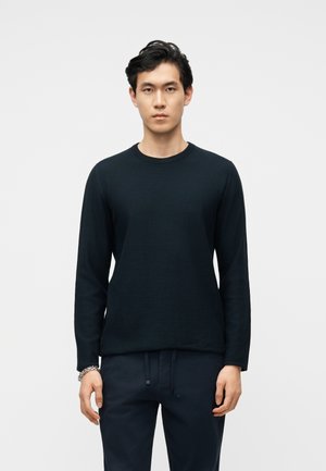 Tânăr care stă în picioare, cu fața înainte, purtând un pulover negru cu mânecă lungă și gât rotund și pantaloni închiși la culoare, cu o brățară argintie la încheietura mâinii stângi.