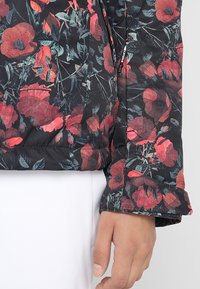 Schwarze Jacke mit floralem Muster aus großen roten Mohnblumen und grünen Blättern, strukturiertem Stoff, mit einer Nahaufnahme des Bündchens.