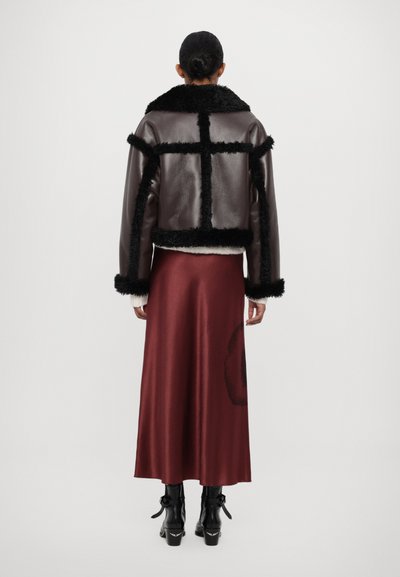 Brown leather jacket with black fur trim, featuring a boxy design and contrasting textured panels, paired with a sleek maroon satin skirt.