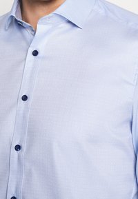 Light blue shirt with a subtle dotted pattern, featuring a pointed collar and navy buttons that contrast against the fabric.