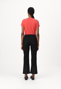 Korallefarbendes Baumwoll-T-Shirt kombiniert mit schwarzen, ausgestellten Jeans, die am Saum ein strukturiertes Muster aufweisen. Das Modell steht mit dem Rücken zur Kamera.