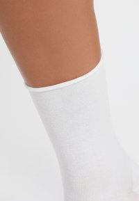 Calcetín de algodón blanco con textura acanalada, que presenta un puerto elástico suave en la parte superior. La imagen muestra el calcetín a la altura del tobillo sobre la piel.