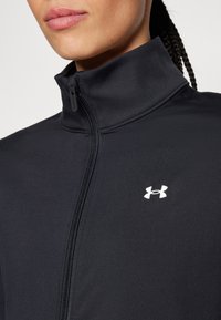 Svart zip-up jacka i slätt tyg, med hög krage och en vit logotyp på vänster bröst. Dragkedjan  är synlig.