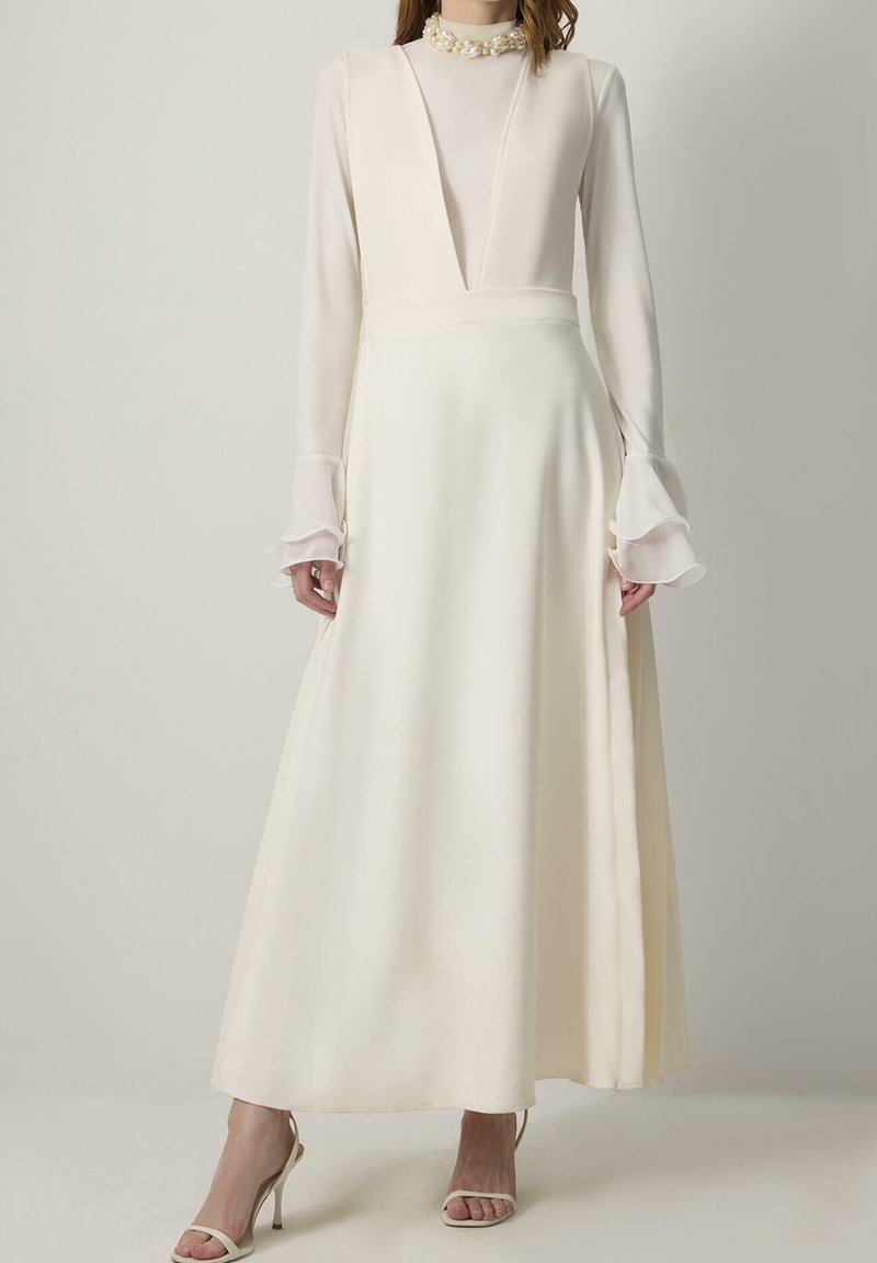 Femme portant une robe blanche à manches longues avec des poignets en tulle volantés et une ouverture devant, associée à des sandales blanches à talons hauts.