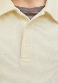Polo crème avec un tissu texturé, doté d'une patte de boutonnage à trois boutons et d'un col. En dessous se trouve un t-shirt col rond marron.