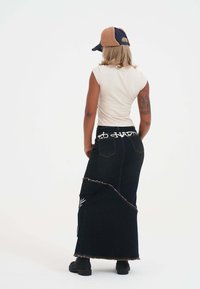 Zwarte denim maxirok met rafelige zoom en contrasterende stiksels, voorzien van een wit logo op de tailleband. Draag het met een beige en marineblauwe pet.