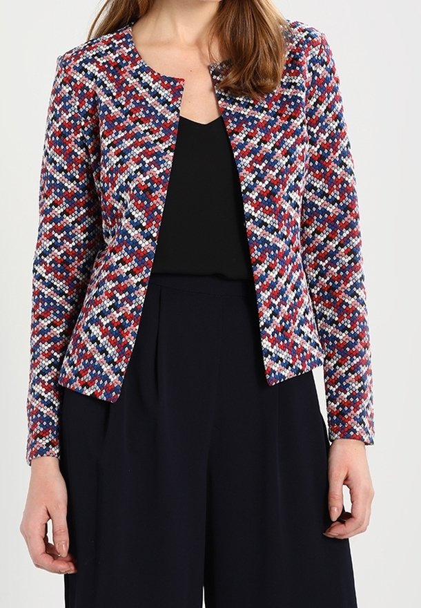 Femme portant une veste cintrée texturée multicolore avec des motifs rouges, bleus, blancs et noirs sur un haut noir et un pantalon noir.
