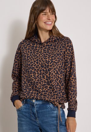 Smilende kvinde iført en brun og sort leopardmønstret hættetrøje med blå jeans, hænderne i lommerne, kigger til siden mod en ensfarvet baggrund.