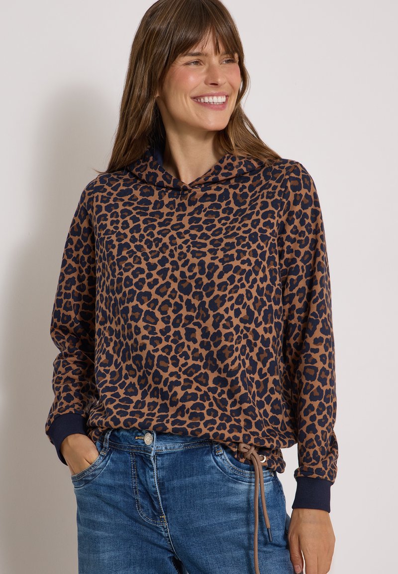 Femme souriante portant un sweat à capuche marron et noir à imprimé léopard avec un jean bleu, mains dans les poches, regardant sur le côté sur un fond uni.
