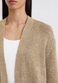 Gebreide cardigan in lichtbeige, met een gestructureerde weving, open voorkant en zachte stof, gedragen over een witte tanktop.