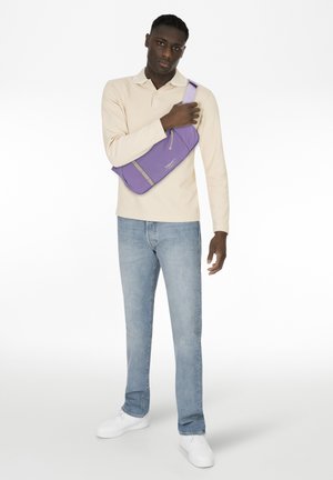 Mann mit dunkler Hautfarbe, der ein beigefarbenes Langarmshirt, hellblaue Jeans, weiße Turnschuhe und eine lila Umhängetasche in einer neutralen Pose trägt.