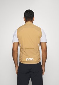 Bež bezrukavni prsluk izrađen od laganog materijala, s džepom na leđima i bijelim "poc" logom na rubu.