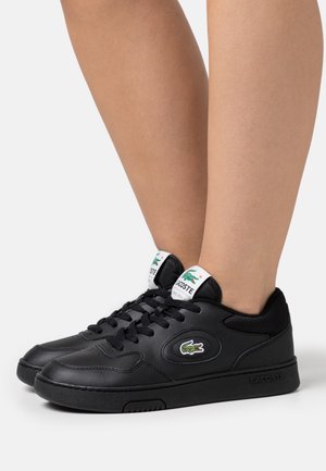Zapatillas negras Lacoste con etiquetas blancas en la lengüeta y logotipo de cocodrilo verde, usadas en los pies sobre un fondo claro y liso.
