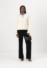 Cardigan blanc en peluche avec des boutons dorés et deux poches avant, associé à un pantalon noir à jambes larges et un sac à main noir.