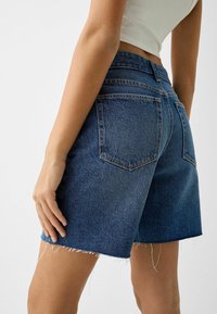 Pantalones cortos de mezclilla en lavado oscuro con dobladillo sin rematar y bordes deshilachados. Presentan bolsillos traseros y detalles clásicos de costura. Ajuste estándar y diseño de talle medio.