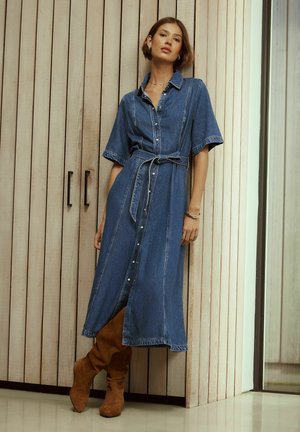 Kobieta opierająca się o drewnianą ścianę, ubrana w dżinsową sukienkę midi z paskiem i brązowe zamszowe kozaki za kolano.