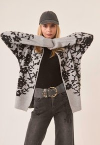 Cardigan gris de gran tamaño con patrones florales negros, top negro de canalé, cinturón gris con acentos de tachuelas y jeans oscuros con detalles bordados.