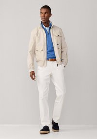 Veste légère beige avec deux poches avant, pull bleu, pantalon blanc, et espadrilles noires à semelle en jute. Fond uni.