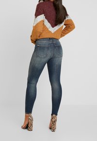 Těsné džíny v tmavě modrém denimovém provedení s vybledlými detaily, oblečené s pulovrem s barevným šikmým vzorem v teplých tónech a s hadími podpatky.