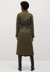 Lang olijfgroen coat met een ceintuur in de taille, oversized kraag en lange mouwen, gemaakt van een zachte stof. Draag het met zwarte knielaarzen.